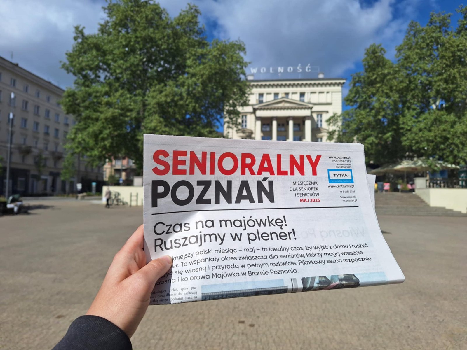 Majowy Senioralny Poznań Ktoś trzyma złożoną gazetę. W tle zabytkowy budynek Arkadii