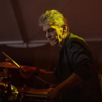 Mężczyzna w średnim wieku, z siwymi włosami i kolczykami w uchu. Ubrany w czarną koszulę z podwiniętymi rękawami. Mężczyzna gra na perkusji.