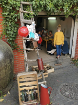 Grupa dzieci majsterkuje w garażu. Pomaga im kobieta. Dookoła leżą rożne urządzenia i narzędzia do majsterkowania.