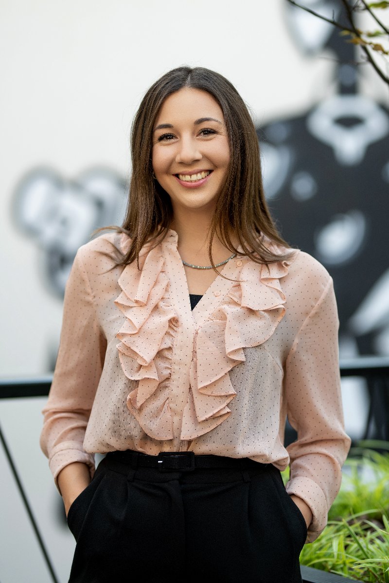 Uśmiechnięta kobieta w pastelowo-różowej koszuli i czarnych spodniach. W jej tle widać rozmazany fragment czarno-białego muralu.
