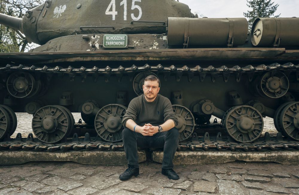 Krzysztof Jankowiak, fot. Grzegorz Dembiński Mężczyzna w dżinsach i ciemnozielonym swetrze siedzi na gąsienicy zabytkowego czołgu zaplatając dłonie. Ma skupiony wyraz twarzy.