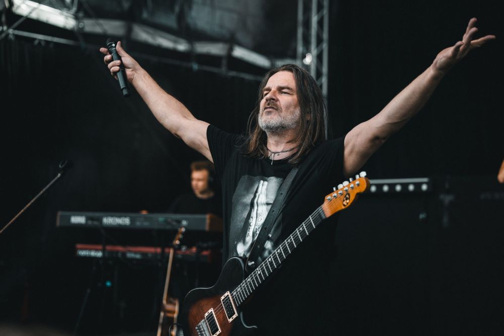 Mężczyzna z gitarą na scenie szeroko rozkłada ręce.