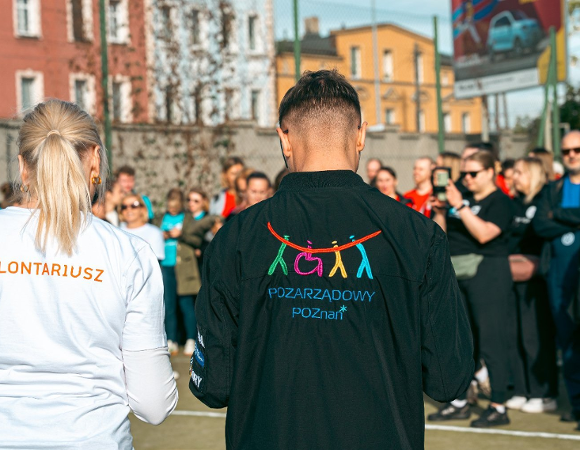 Na zdjęciu dużo ludzi, dwie osoby stoją tyłem do obiektywu, mają koszulki z napisem wolontariusz