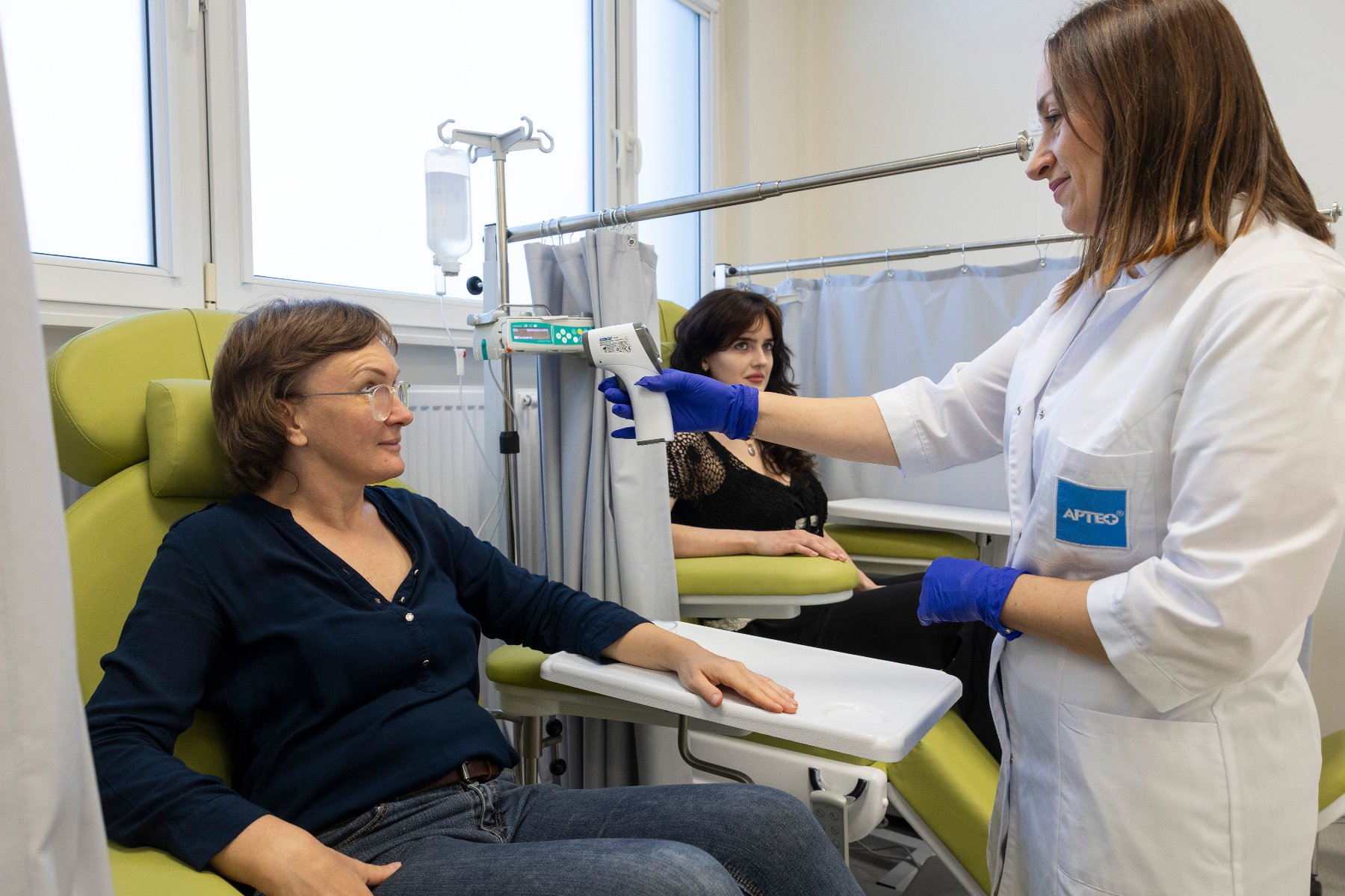 Na zdjęciu dwie kobiety siedzące na fotelach lekarskich, obok nich stoi kobieta w białym fartuchu z termometrem w dłoni