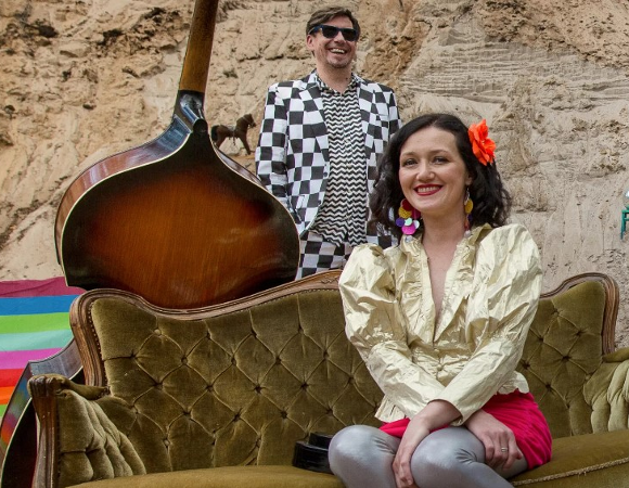 Na zdjęciu kobieta siedząca na kanapie, za nią stoi mężczyzna, obok duży instrument smyczkowy