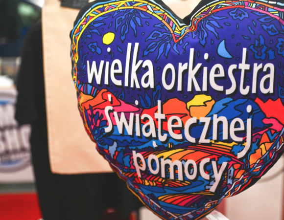 Na zdjęciu poduszka w kształcie kolorowego serca z napisem: "Wielka Orkiestra Świątecznej Pomocy". Tło rozmazane.