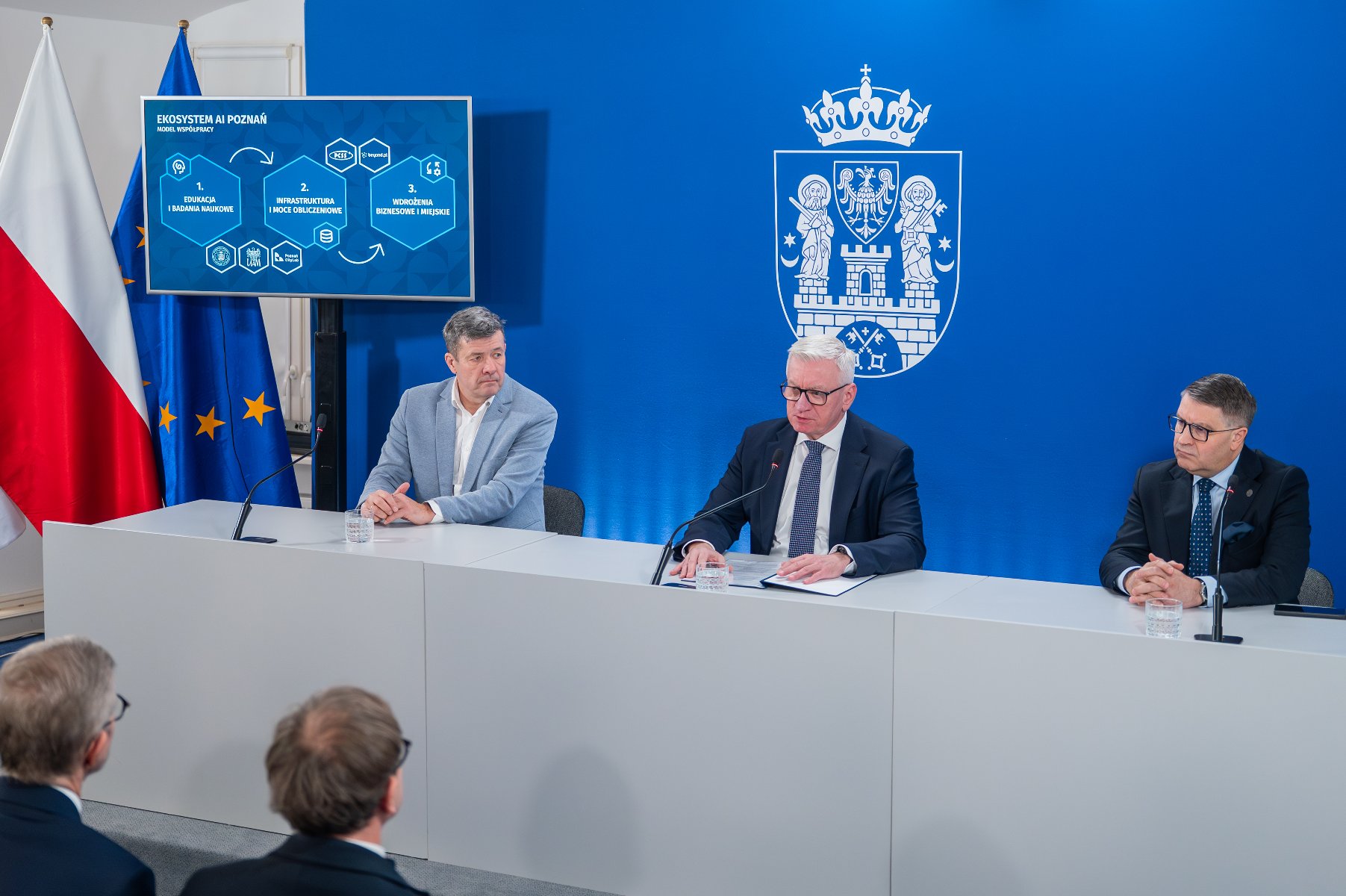 Na zdjęciu trzej mężczyźni w garniturach, siedzący za stołem podczas konferencji prasowej