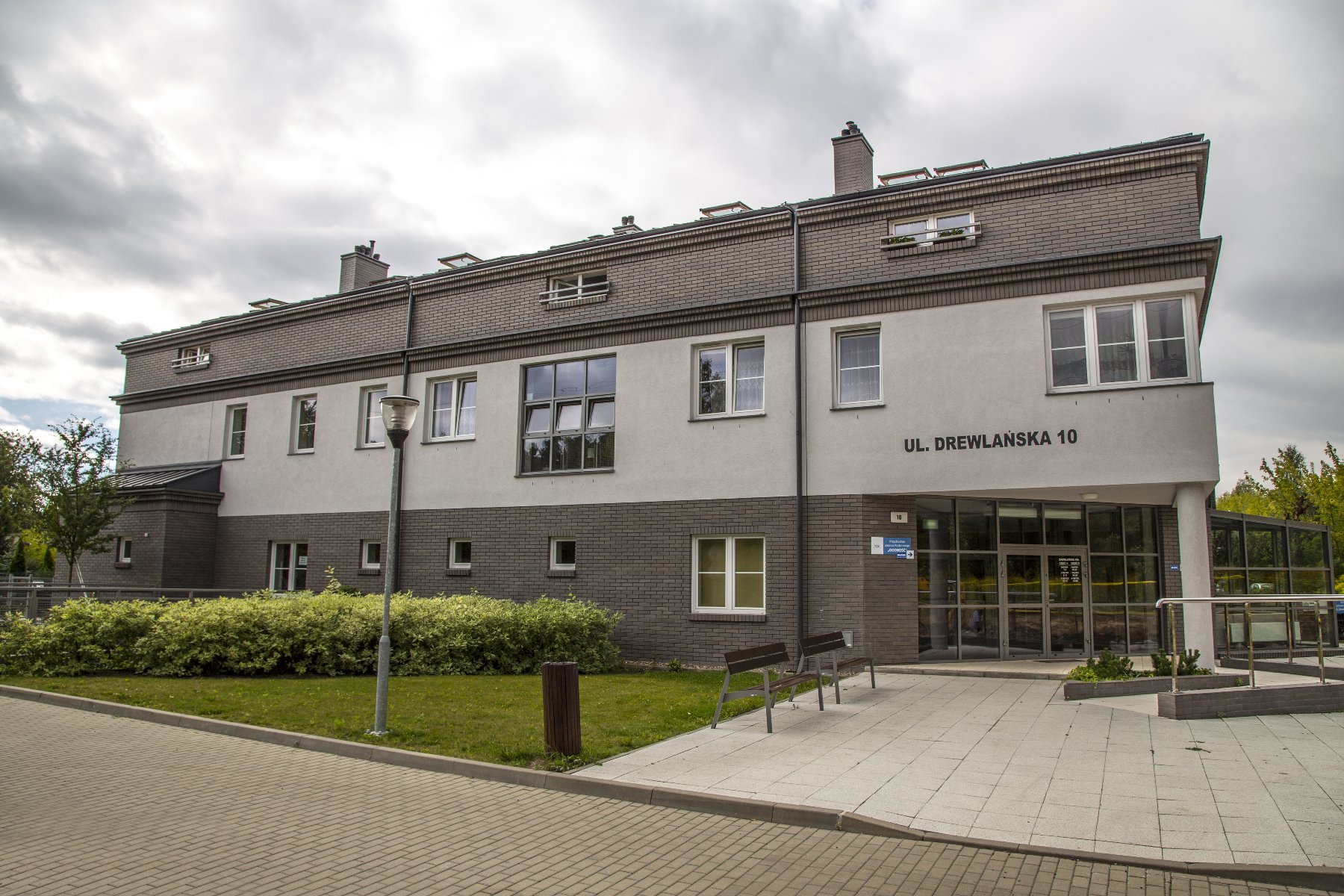 Na zdjęciu widać budynek przy ul. Drewlańskiej 10. Budynek ma 3 kondygnacje. Z prawej strony widać wejście do budynku. Jest przeszklone. Przed budynkiem znajduje się trawnik, chodnik i ławeczki.