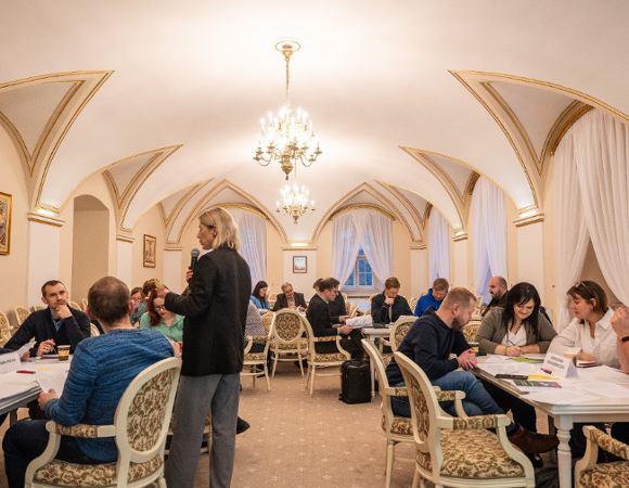 Na zdjęciu widać dużą grupę osób, które siedzą przy kilku stolikach w dużej, eleganckiej sali Urzędu Miasta.