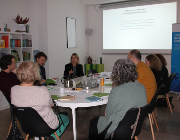 Na zdjęciu widać grupę osób siedzących przy owalny, dużym białym stole. Na stole znajdują się pomoce warsztatowe. W tle widać rzutnik z prezentacją.