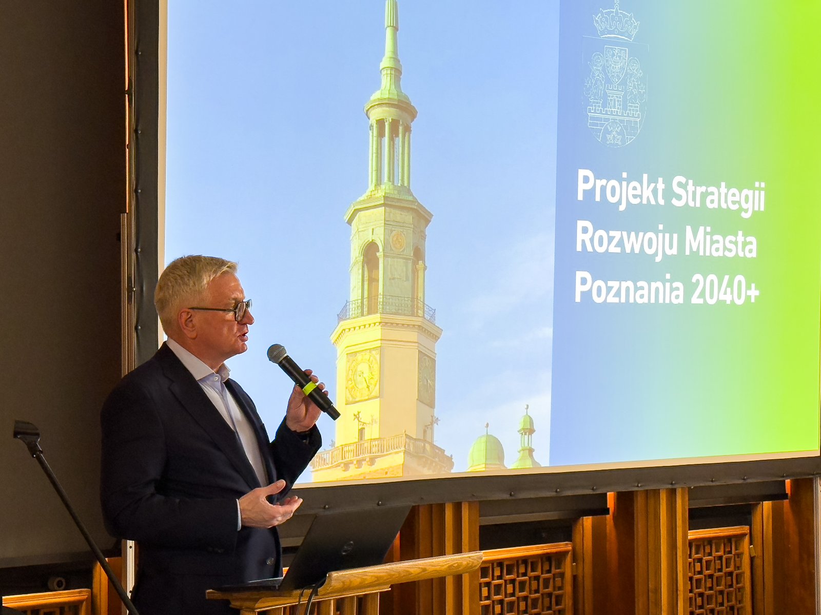 Uroczyste otwarte spotkania przez pana prezydenta Jacka Jaśkowiaka na zdjęciu widac pana prezydenta miasta Poznania Jacka Jaśkowiaka, który w ręce trzyma mikrofon i przemawia. Za nim widać prezentację wyświetalną na projektorze, na której widać ratusz poznański oraz napis "Projekt Strategii Rozwoju Miasta Poznania 2040+"