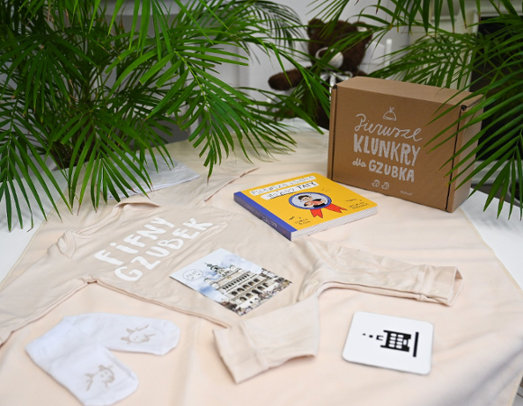 Na zdjęciu wyprawka: na stoliku rozłożone śpioszki i inne elementy wyprawki, w tle widać rośliny