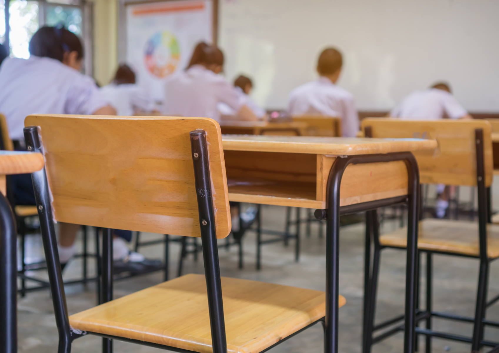 Na zdjęciu znajduje się klasa szkolna, na pierwszym planie jest puste drewniane krzesło przy drewnianej ławce, w dalszej perspektywie znajdują się niewyraźne sylwetki uczniów ubranych w białe koszulki.