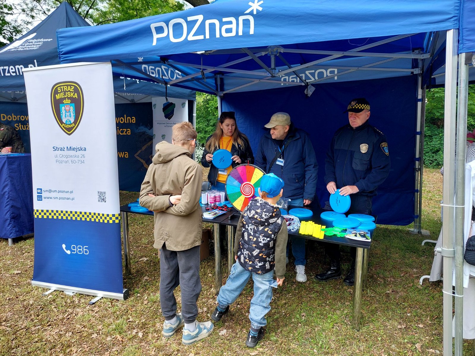 Stoisko profilaktyki SMMP Na zdjęciu znajduje się niebieski namiot z logo Poznania, 3 osoby ze straży miejskiej, 2 kobiety i mężczyzna prezentują materiały promocyjne przy stole, na stole jest kolorowe koło do kręcenia i losowania zadań, przed namiotem stoi dwóch chłopców