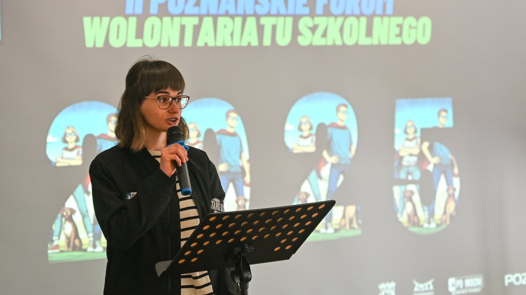 Na zdjęciu młoda kobieta, mówiąca coś do mikrofony, w tle ekran