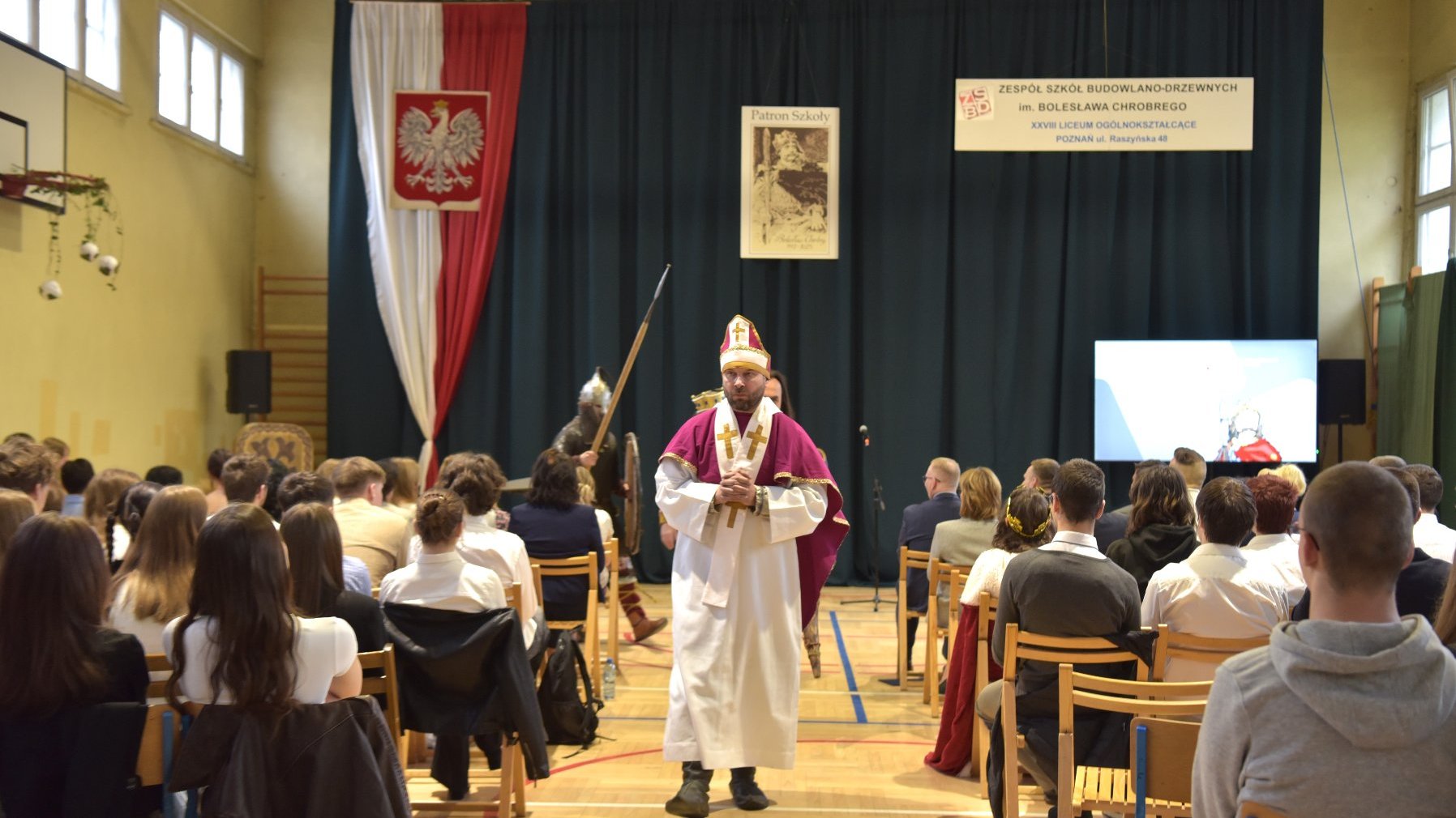 Zdjęcie przedstawia uczniów siedzących na sali gimnastycznej oraz wychodzących z sali mężczyzn w przebraniach średniowiecznych.