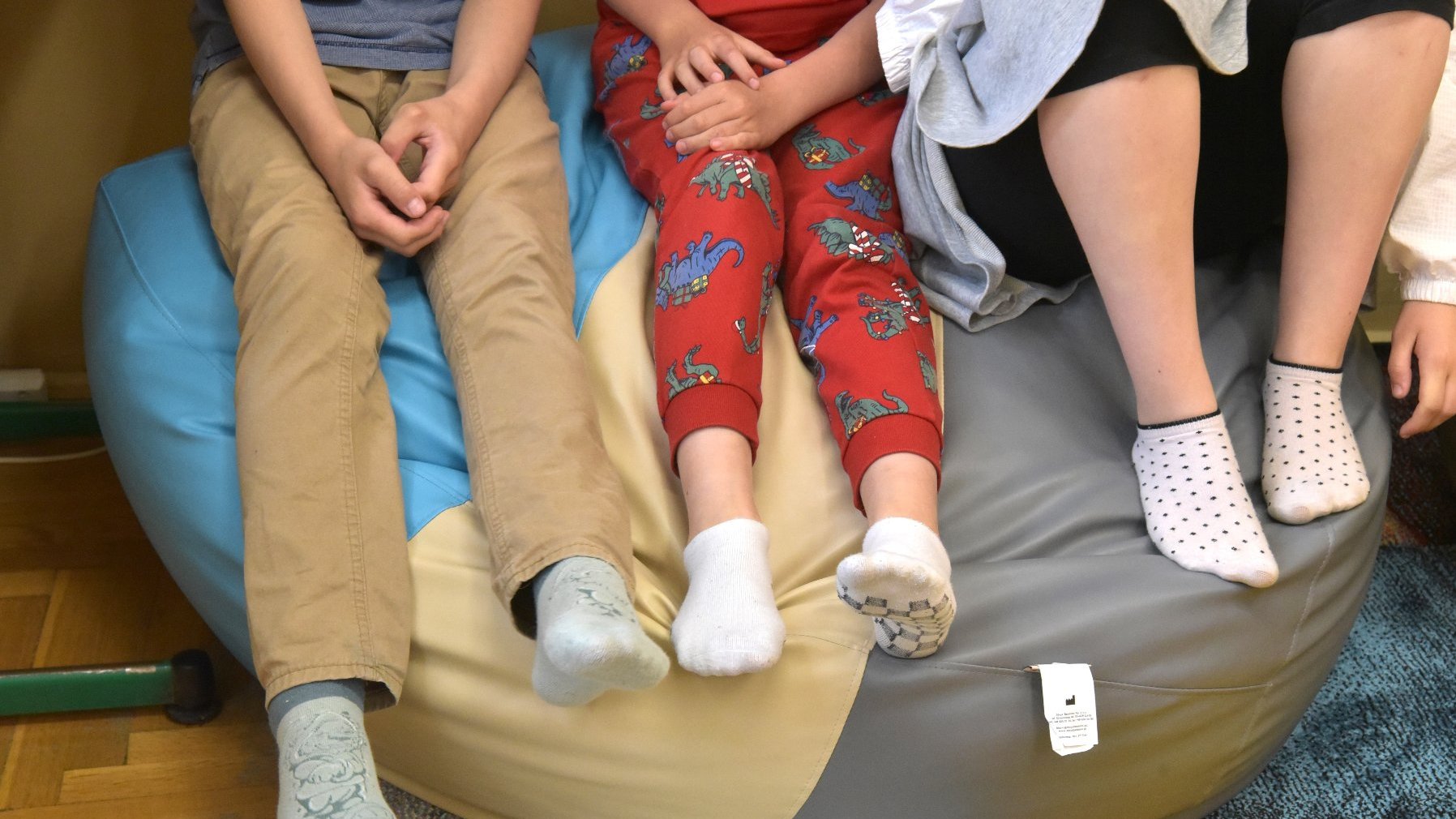 Zdjęcie przedstawia dzieci siedzące na pufie. Widać tylko ich nogi.
