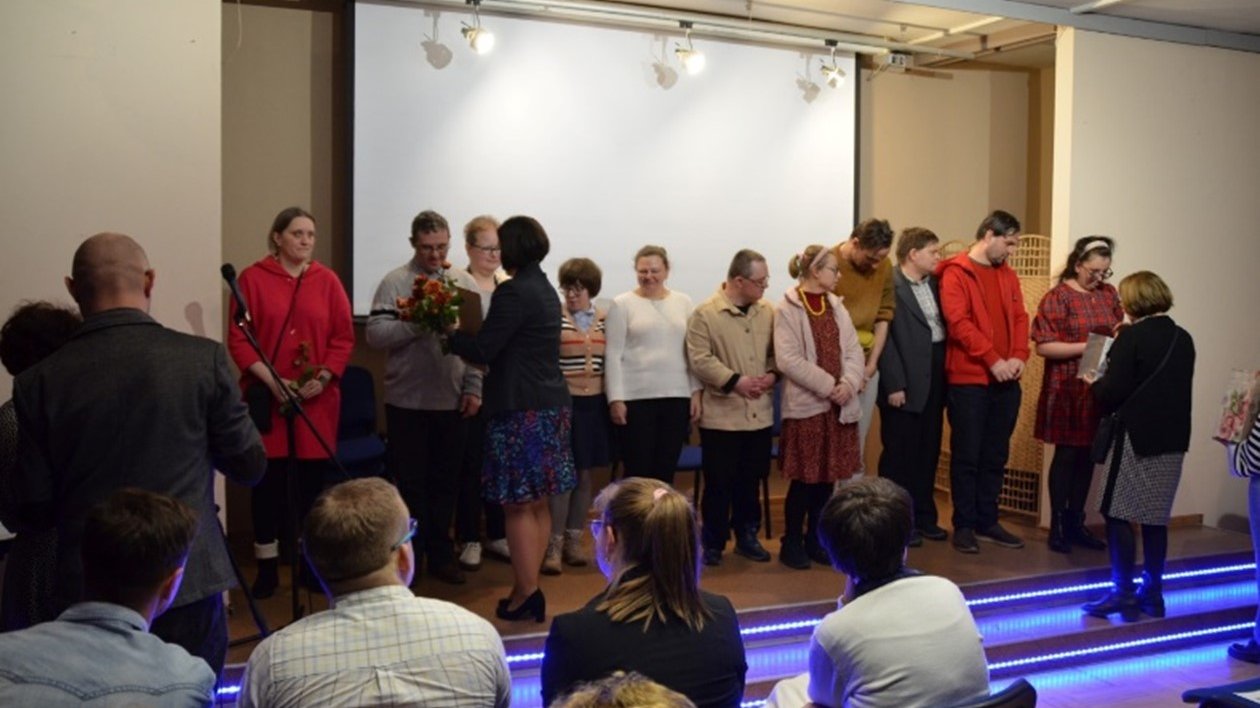 Liczna grupa osób stoi na scenie. Dwie kobiety wręczają dyplomy i kwiaty autorom wierszy.