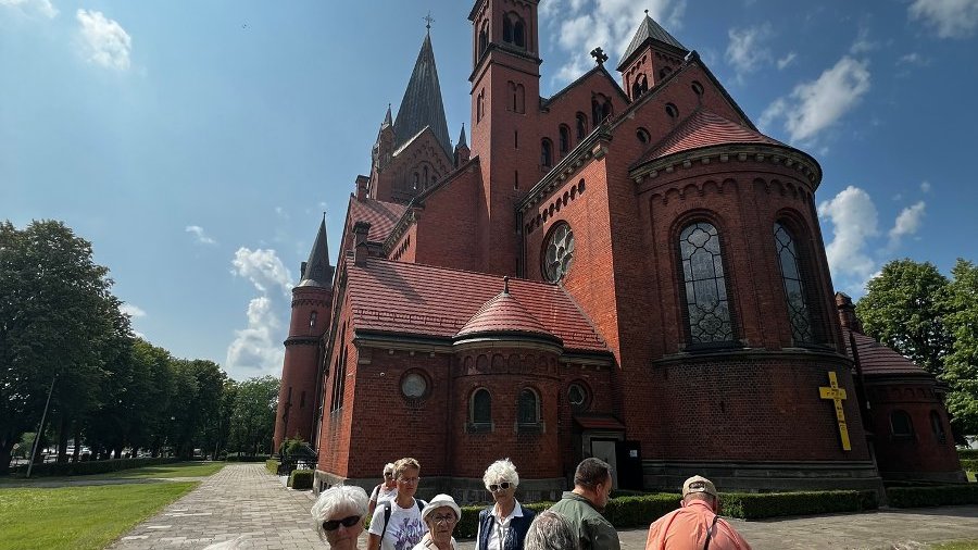 Grupa seniorów przy Kościele Zwiastowania Najświętszej Maryi Pannie. Kościół jest duży, zbudowany z czerwonej cegły.