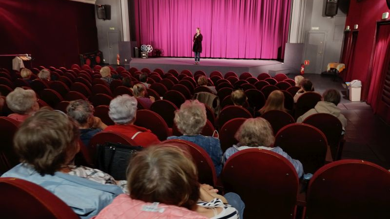 Grupa osób siedzi na sali kinowej słuchając osoby zapowiadającej film.