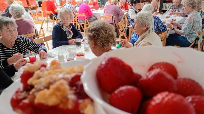 Na pierwszym planie talerzyk z ciastem i miska z truskawkami, w tle grupa seniorów przy stołach w czasie posiłku.