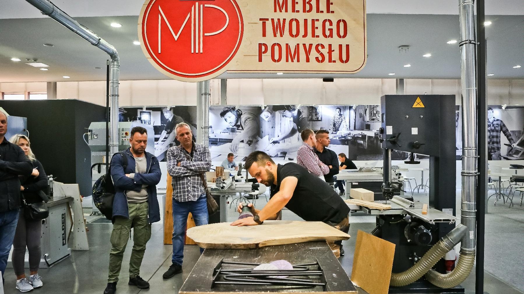 Mężczyzna pracuje na dużym kawałku drewna. Jego pracę obserwuje grupka osób. Nad nim wisi napis "Meble Twojego Pomysłu". Wszystko dzieje się we wnętrzu hali targowej.