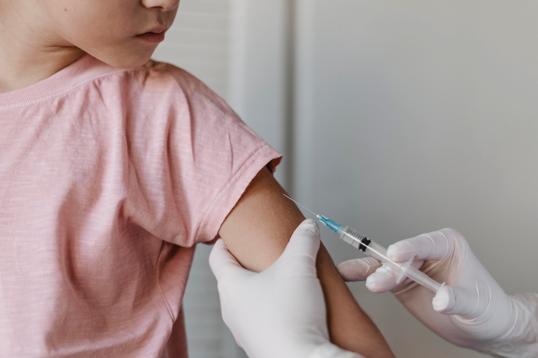 Na zdjęciu dziecko w różowej koszulce podczas szczepienia - grafika artykułu