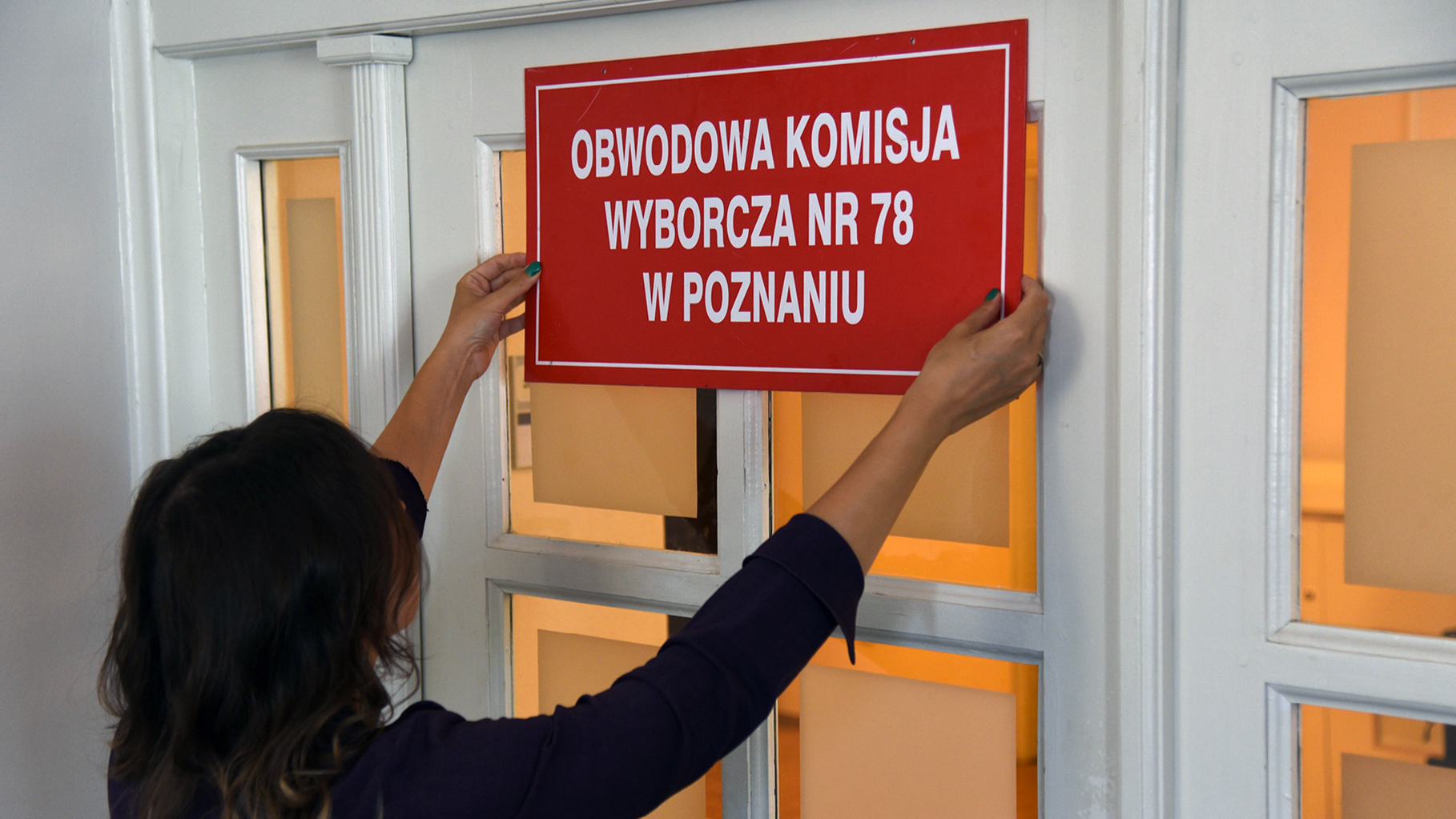 Osoba wiesza na drzwiach tablicę z napisem "Obwodowa komisja wyborcza nr 78 w Poznaniu" - grafika artykułu