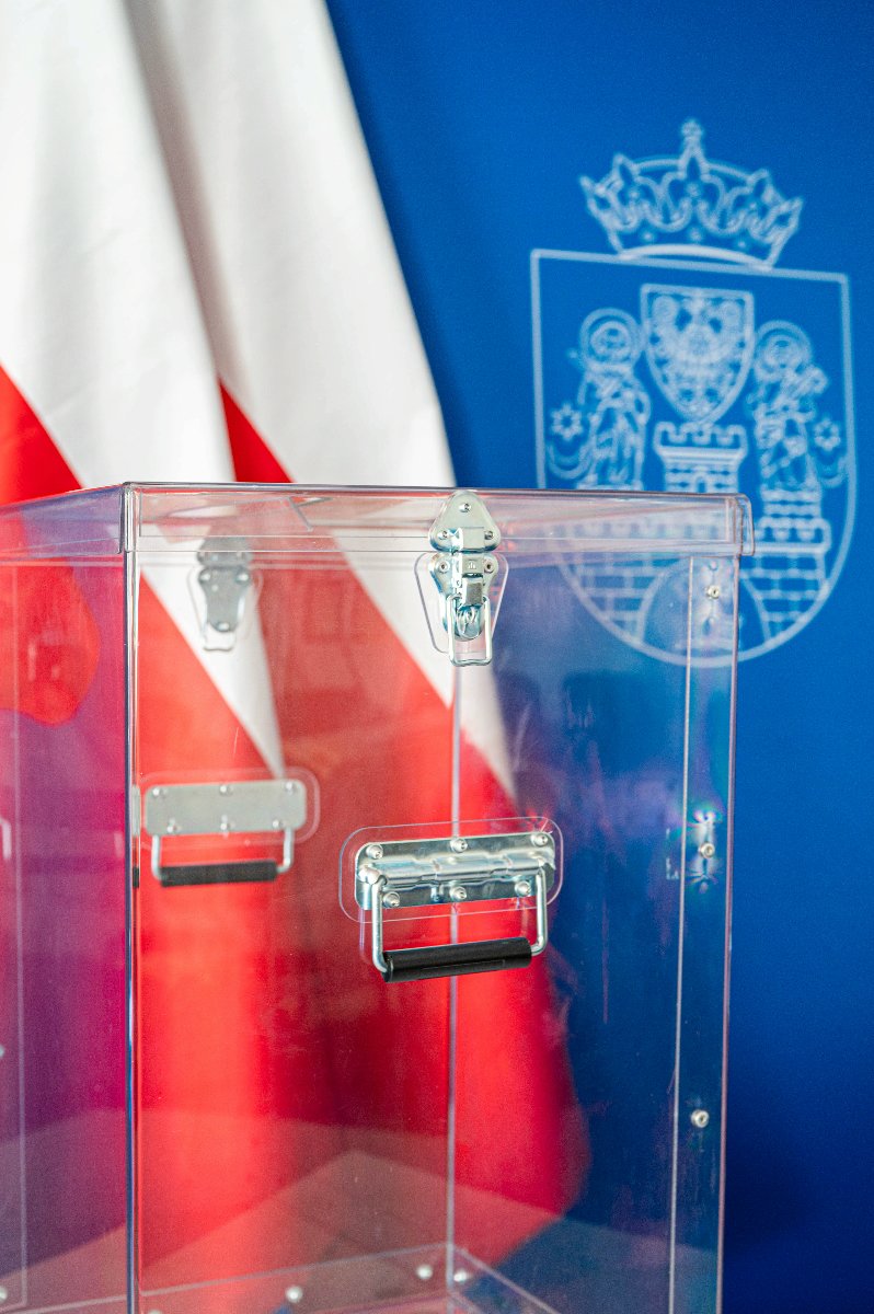 Przeźroczysta urna wyborcza, w tle dwie flagi biało-czerwone i herb Miasta Poznania na niebieskim tle - grafika artykułu