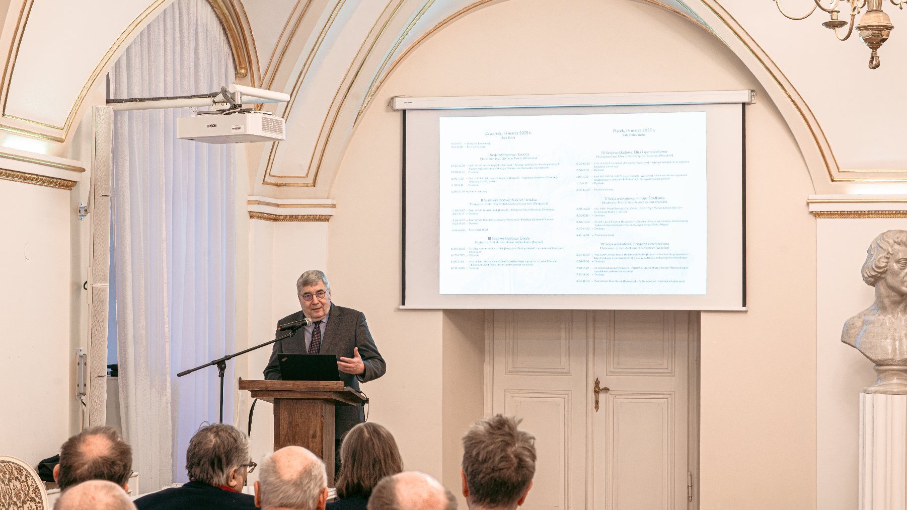 Zdjęcie przedstawia przewodniczącego rady miasta mówiącego do mikrofonu na mównicy oraz tłum siedzących ludzi na sali.