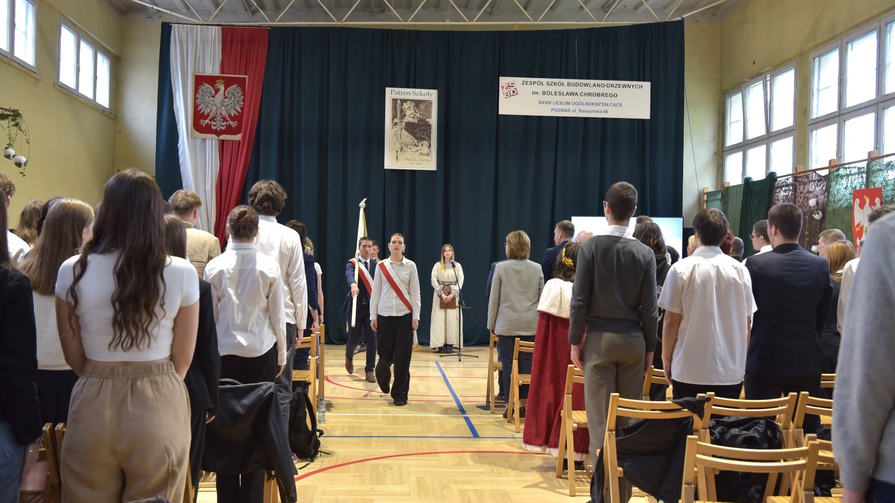 Zdjęcie przedstawia uczniów stojących na sali gimnastycznej. Trwa wyprowadzanie sztandaru.