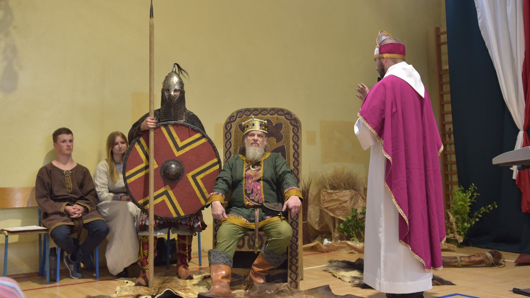 Zdjęcie przedstawia trzech mężczyzn z przebraniach średniowiecznych. Król siedzi na tronie, biskup zakłada mu koronę, obok stoi wojownik.