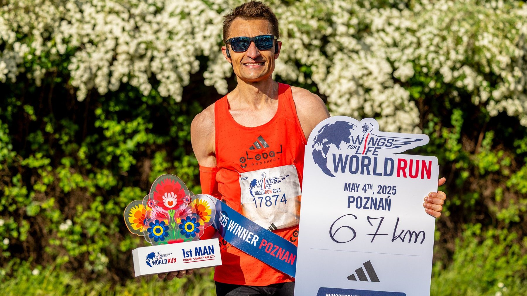 Dariusz Nożyński zwycięzcą biegu Wings for Life World Run w Poznaniu, fot. Damian Kramski - mężczyzna stoi w czerownym stroju i okularach przeciwsłonecznych na tle zieleni i trzyma nagrodę za zwycięstwo w zawodach