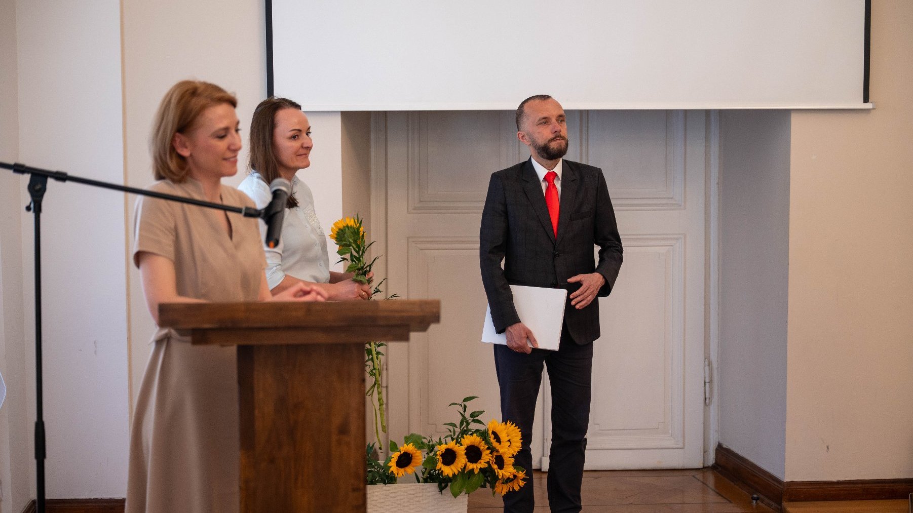 Trzy osoby stoją w formalnym otoczeniu. Jedna osoba przemawia za drewnianym podium z mikrofonem, podczas gdy druga trzyma bukiet słoneczników. Trzecia osoba, ubrana w garnitur z czerwoną krawatą, trzyma teczkę i stoi przed białymi drzwiami.