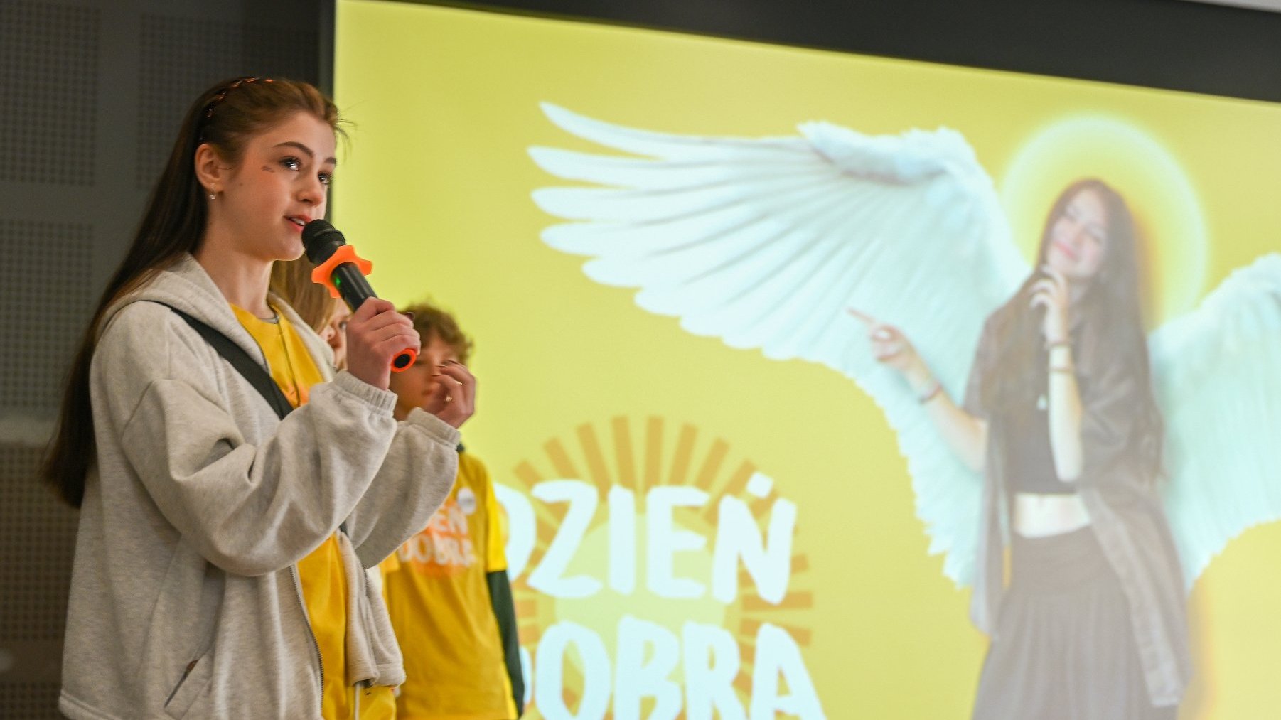 Na zdjęciu młoda dziewczyna, w tle ekran, na nim postać ze skrzydłami