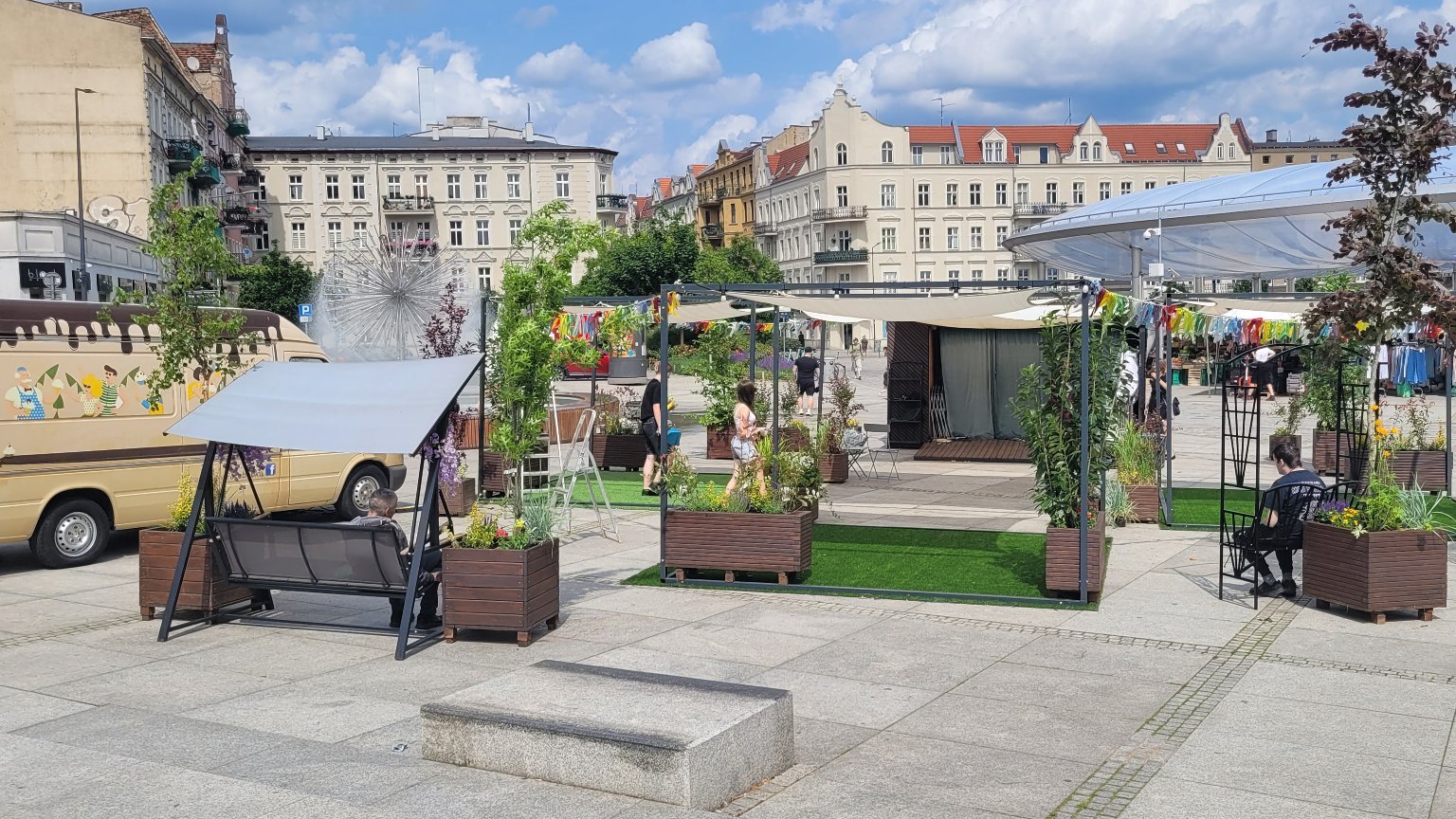 Widok na osoby które rozkładają strefę Łazarskiego Lata. Po lewej stronie bus monterów. Widać rozłożone huśtawki stoliki, baldahim i kolorowe wstążki, które powiewają zawieszone wokół strefy.