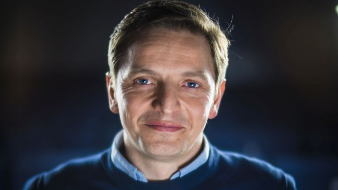 Dyrektor teatru uśmiecha się subtelnie i patrzy prosto w obiektyw, ma na sobie elegancki ciemny sweter, spod którego wystaje błękitny kołnierz koszuli.