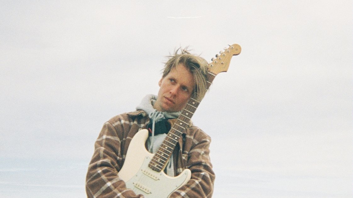 Mężczyzna trzyma gitarę i opiera o nią głowę. W tle jezioro.