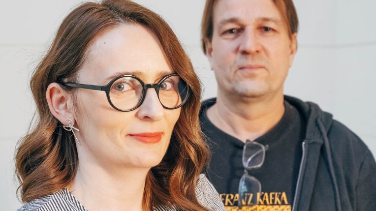 Kobieta w okularach z czarną oprawką i ustami pomalowanymi na czerwono stoi z przodu, uśmiecha się do obiektywu. Z tyłu mężczyzna w bluzie i T-shircie.
