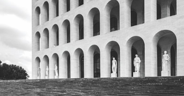 Wejście do jednej z włoskich budowli, wyglądającej bardzo nowocześnie - na ścianach jest mnóstwo otworów okiennych w tych samych kształtach. Na ziemi, przed wejściami, stoi kilka marmurowych rzeźb.