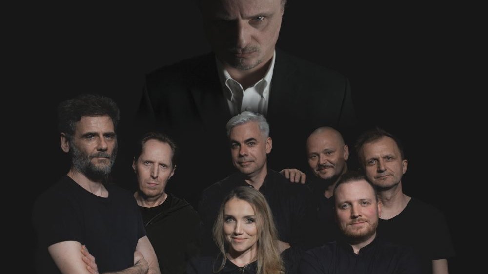 Grupowe zdjęcie członków teatru. Wszyscy ubrani są na czarno, pozują na czarnym tle. Z tyłu większa sylwetka mężczyzny, pod nią pozostałe osoby.