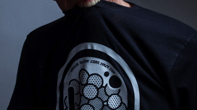 Łysy mężczyzna w okularach przeciwsłonecznych chwyta zębami swój czarny T-shirt z nadrukiem.