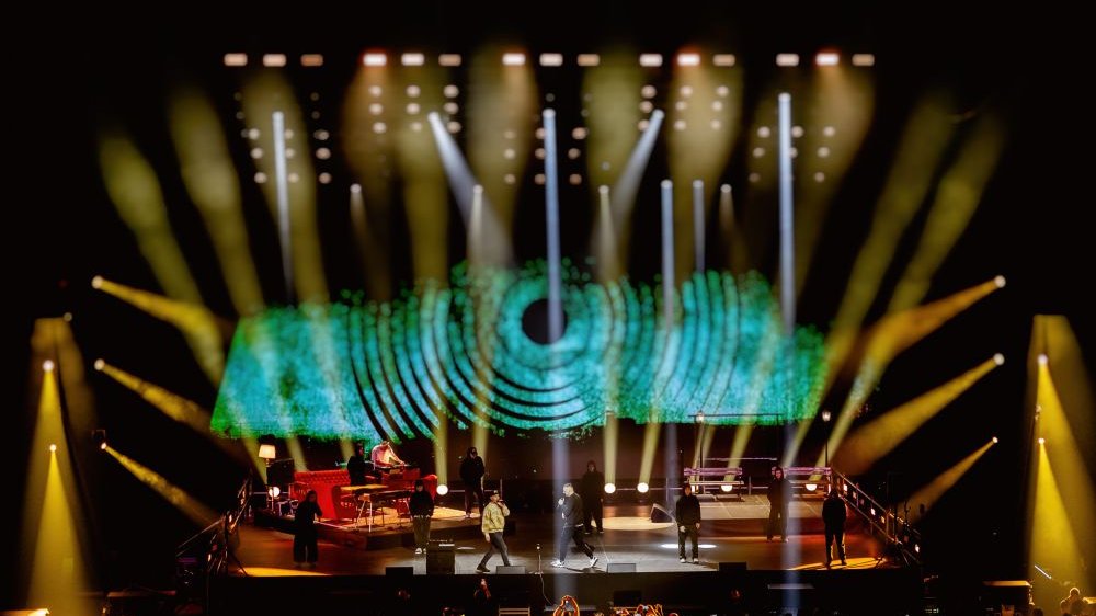 Scena oświetlona ostrym, turkusowym światłem, występują na niej artyści. Tuż przed sceną stoją fani z wyciągniętymi wysoko ramionami, niektórzy filmują telefonem.