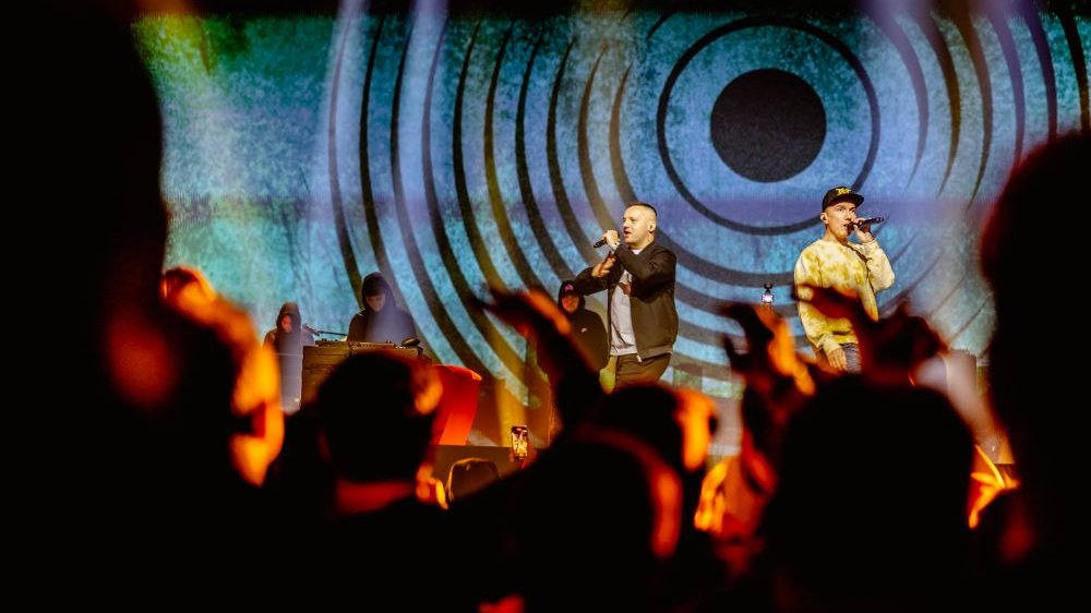 Artyści rapują na scenie, tłum fanów niedaleko sceny wyciąga wysoko ręce.