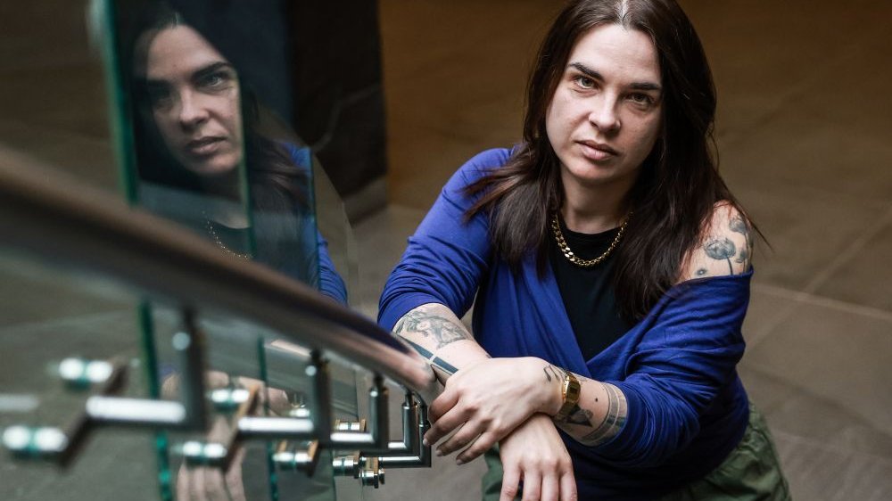 Tłumaczka trzyma ręce na balustradzie schodów i patrzy na fotografa, który znajduje się kilka schodków wyżej. Widać jej ramiona pokryte tatuażami.
