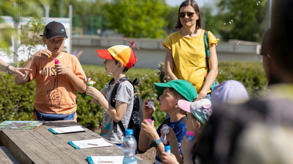 Przy stoliku w plenerze stoi kilkoro dzieci, głównie chłopcy, w kolorowych czapkach z daszkeim. Dmuchają bańki. Za nimi kobieta w żółtej koszulce i okularach słonecznych.