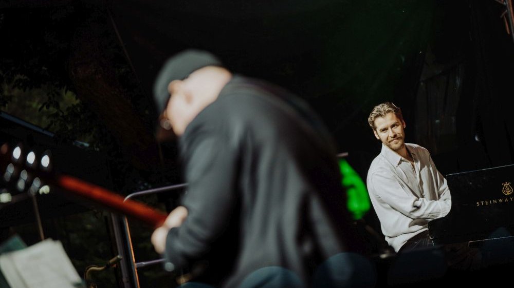 Pianista w białej koszuli uśmiecha się patrząc w stronę basisty podczas występu.