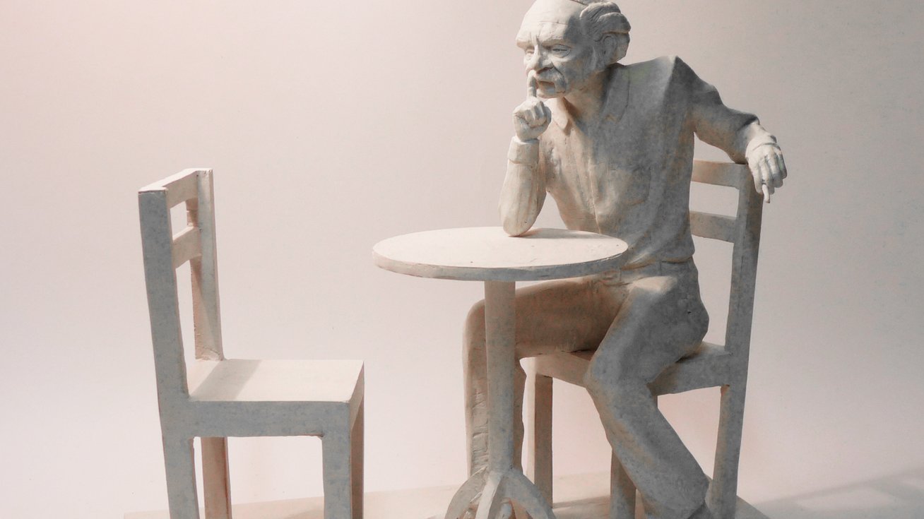 Projekt: Bohdan Smoleń siedzący na krześle przy niewielkim stoliku, zamyślony. Prawy łokieć opiera na stole i dłonią dotyka brody, lewa dłoń na oparciu krzesła. Naprzeciw drugie, puste krzesło - grafika artykułu