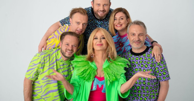 Zdjęcie piosenkarki Majki Jeżowskiej, w jaskrawej, zielonej kurtce. Artystka otoczona jest czterema mężczyznami i jedną kobietą, każdy z nich ubrany jest w kolorowy t-shirt..