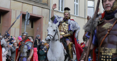Zdjęcie korowodu na ulicy święty Marcin, na białym koniu siedzi mężczyzna przebrany za świętego Marcina, w stroju legionisty, który macha ręką do ludzi. W tle osoby oglądające paradę.